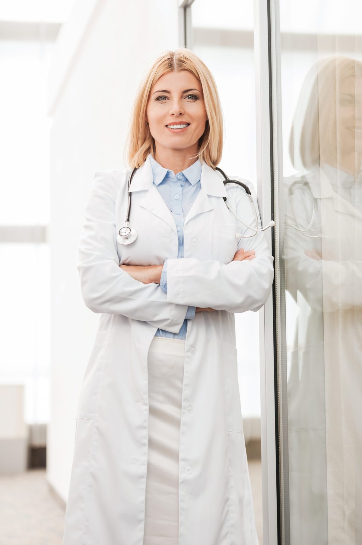 Confident female doctor. Confident female doctor in white uniform looking at camera and smiling while leaning at the glass wall